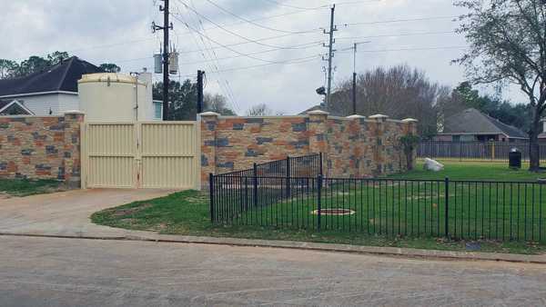 Stone wall with decorative pillars and a tan metal gate in front of a water storage tank. A black metal fence borders a grassy area under a cloudy sky near power lines.